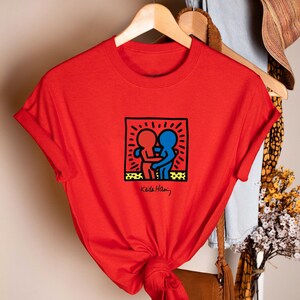 May include: A red t-shirt featuring a Keith Haring design of two embracing figures in red and blue, within a black outlined square. The artist's signature is below the graphic. The shirt is hanging on a wooden hanger.