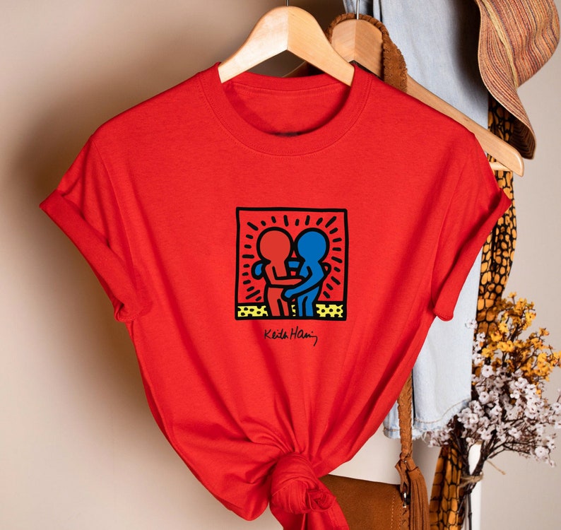 May include: A red t-shirt featuring a Keith Haring design of two embracing figures in red and blue, within a black outlined square. The artist's signature is below the graphic. The shirt is hanging on a wooden hanger.