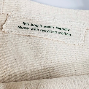 Op de afbeelding: Close-up van een beige canvas draagtas. Een wit rechthoekig label is op de tas genaaid, met de tekst "This bag is earth friendly Made with recycled cotton" in groen gedrukt. De tas is gemaakt van een textiel met een natuurlijke kleur.