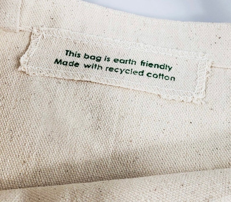 Op de afbeelding: Close-up van een beige canvas draagtas. Een wit rechthoekig label is op de tas genaaid, met de tekst "This bag is earth friendly Made with recycled cotton" in groen gedrukt. De tas is gemaakt van een textiel met een natuurlijke kleur.