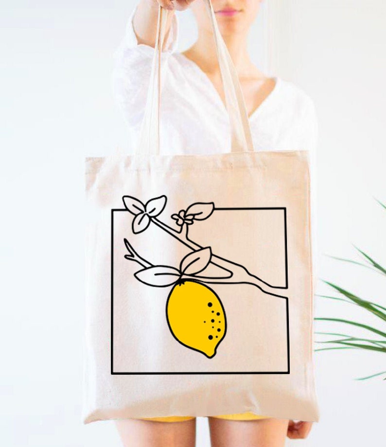 May include: Cream-colored tote bag featuring a black and yellow lemon illustration. The design includes a yellow lemon with black dots, leaves, and a branch within a black square. The bag has long handles.