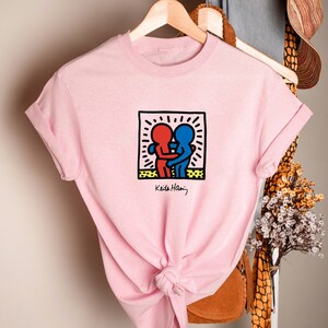 May include: A light pink t-shirt featuring a graphic of two embracing figures in red and blue within a black-outlined square. The design is signed "Keith Haring" in black. The shirt is hanging on a wooden hanger.