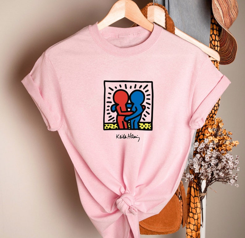 May include: A light pink t-shirt featuring a graphic of two embracing figures in red and blue within a black-outlined square. The design is signed "Keith Haring" in black. The shirt is hanging on a wooden hanger.