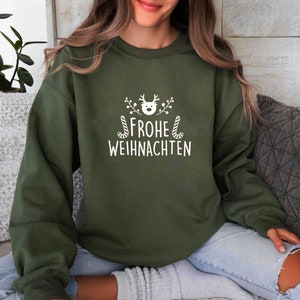 May include: An olive green sweatshirt with the German text "FROHE WEIHNACHTEN" in white, meaning "Merry Christmas." The design includes a reindeer head, sprigs, and candy canes. A cozy, festive garment for the holiday season.