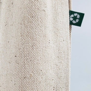 Op de afbeelding: Close-up van een beige canvas draagtas met een getextureerde weefsel. Een klein, donkergroen label met een wit recycling symbool is bevestigd aan de zijkant van de tas. De tas is gemaakt van een duurzaam, milieuvriendelijk materiaal.