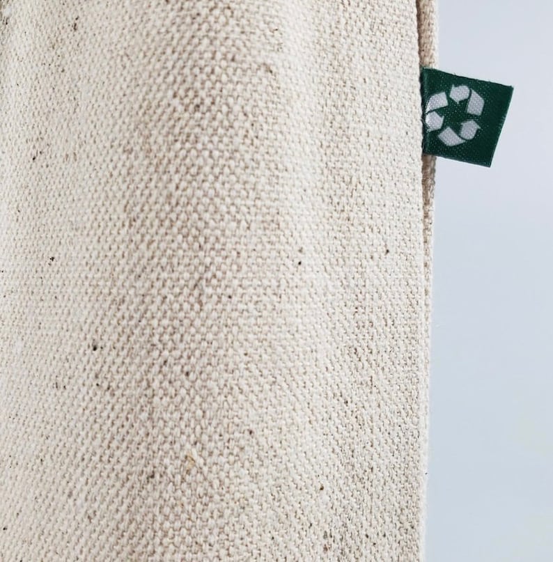 Op de afbeelding: Close-up van een beige canvas draagtas met een getextureerde weefsel. Een klein, donkergroen label met een wit recycling symbool is bevestigd aan de zijkant van de tas. De tas is gemaakt van een duurzaam, milieuvriendelijk materiaal.