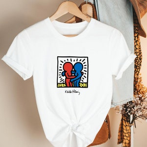 May include: White t-shirt featuring a graphic of two embracing figures in red and blue, within a black-outlined square with a yellow border. The design is signed "Keith Haring" in black. The shirt is hanging on a wooden hanger.
