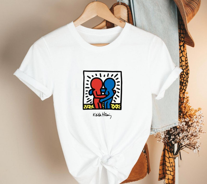 May include: White t-shirt featuring a graphic of two embracing figures in red and blue, within a black-outlined square with a yellow border. The design is signed "Keith Haring" in black. The shirt is hanging on a wooden hanger.
