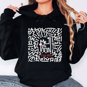 May include: Black hoodie featuring a white graphic of a cat with a heart and abstract lines. The hoodie has the text "Keith Haring" in red. The cat design is centered on the front of the hoodie.