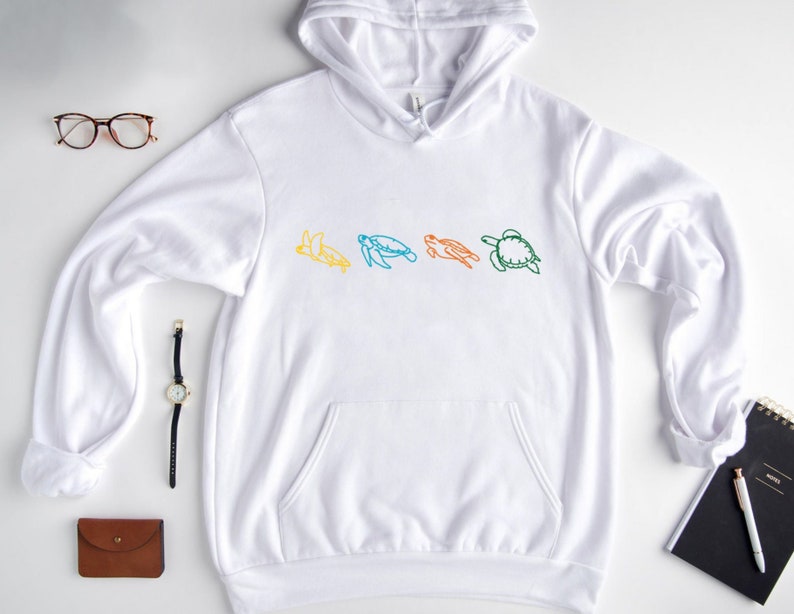 May include: White hoodie with a line drawing of four sea turtles in different colors: yellow, blue, green, and orange. The turtles are facing right and are in a line.