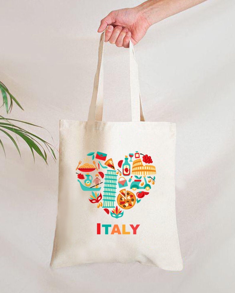 Op de afbeelding: Cr&egrave;mekleurige draagtas met een hartvormig ontwerp gevuld met Italiaanse iconen zoals de scheve toren van Pisa, pizza en het Colosseum. Het woord "ITALY" is in het rood gedrukt. De tas wordt door een hand vastgehouden.