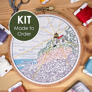 May include: An embroidery kit featuring a circular wooden hoop with a lighthouse landscape design on white fabric, surrounded by colorful embroidery floss spools, metal scissors, and a needle tin with a cactus pattern. Text reads KIT Made to Order.