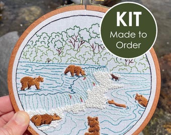 May include: An embroidery hoop containing a fabric panel with a stitched scene of brown bears in a river with a waterfall. The design features green trees, blue water, and white French knots for foam. A circular overlay displays the text KIT Made to Order.