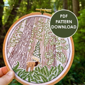 May include: An embroidery hoop containing a fabric panel with a stitched forest scene featuring tall redwood trees, green ferns, and an elk. The design includes a circular overlay with the white text PDF PATTERN DOWNLOAD on a dark green background.