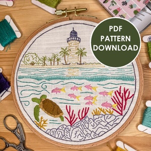May include: Embroidered hoop art featuring a lighthouse on a small island with palm trees, a turtle swimming in the ocean, coral reefs, and colorful fish. The hoop is surrounded by embroidery floss spools and a small tin of embroidery needles. The text "PDF PATTERN DOWNLOAD" is displayed on a green circle.