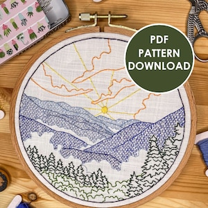 May include: A completed embroidery hoop featuring a mountain landscape design. The design includes a sun, blue mountains, green trees, and orange and yellow clouds. The hoop is on a wooden surface with embroidery floss in various colours.