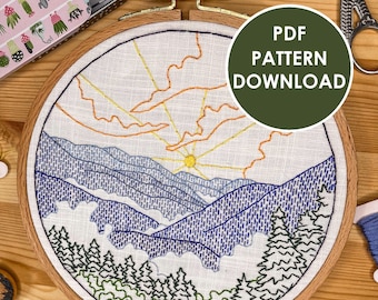May include: A finished embroidery hoop with a mountain landscape design. The design features a sun, blue mountains, green trees, and orange and yellow clouds. The hoop is on a wooden surface with embroidery floss in various colors.