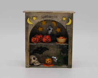 May include: A miniature Halloween-themed display cabinet. The cabinet is made of wood and features an arched opening with a shelf. Inside are miniature pumpkins, a ghost, a bat, a skull, and other Halloween decorations. The cabinet is decorated with yellow moons and stars.