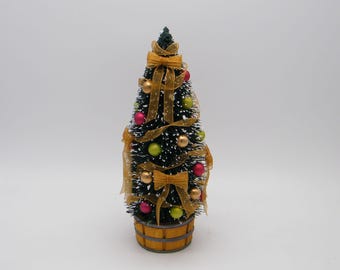 May include: A miniature Christmas tree with green needles, adorned with gold ribbons, bows, and colorful ornaments. The tree is set in a wooden-style base, creating a festive holiday decoration.