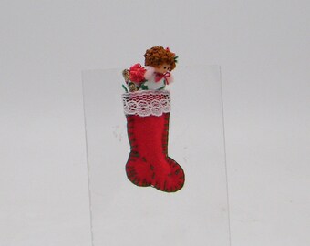 May include: A small red fabric Christmas stocking ornament featuring dark green stitched edges and a white lace trim at the top. A miniature doll with brown curly hair and a white outfit is tucked inside the stocking along with small red floral accents.