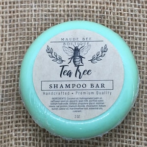 May include: A teal green shampoo bar with a brown label that reads "Maude Bee Boutique Tea Tree Shampoo Bar Handcrafted - Premium Quality" and lists the ingredients. The bar is 3 oz.