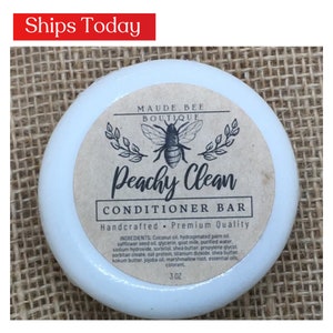 May include: A white round container with a brown label that reads "MAUDE BEE BOUTIQUE Peachy Clean CONDITIONER BAR Handcrafted Premium Quality INGREDIENTS: Coconut oil, hydrogenated palm oil, safflower seed oil, glycerin, goat milk, purified water, sodium hydroxide, sorbitol, shea butter, propylene glycol, sorbitan oleate, oat protein, titanium dioxide, shea butter, kokum butter, jojoba oil, marshmallow root, essential oils, colorant. 3 OZ"