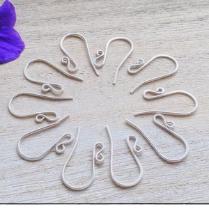 May include: A close-up of ten silver earring hooks arranged in a circle on a light wood surface. The hooks are all the same size and shape, and they are made of a thin, flexible wire.