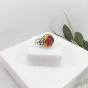 May include: A silver ring with a red gemstone set in a gold bezel. The ring has a detailed floral design.