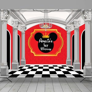 May include: A red and gold birthday backdrop featuring a Mickey Mouse silhouette with a crown, the text "Amelia's 1st Birthday", and a checkered floor. The backdrop has white columns and a chandelier.