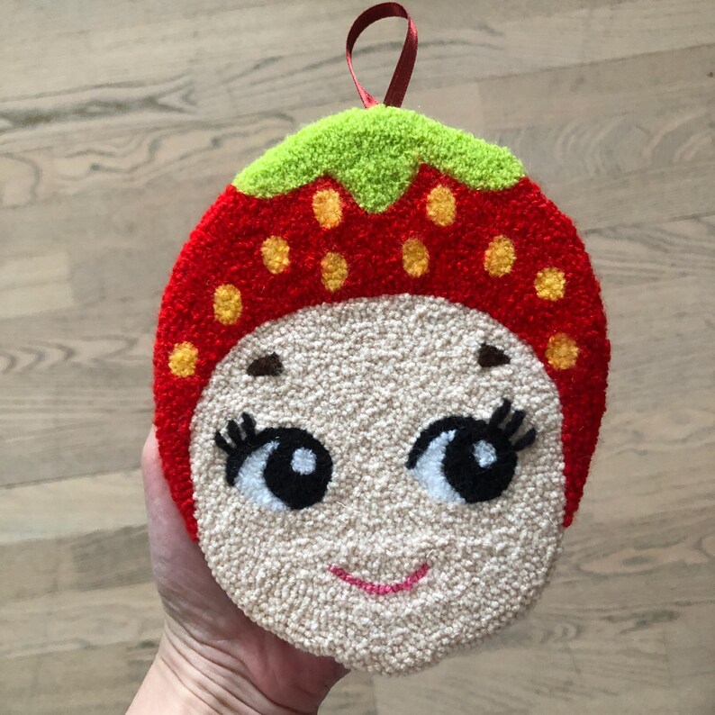 Puede incluir: Una fresa de fieltro rojo y verde con una cara blanca, ojos negros y una sonrisa rosa. La fresa tiene un tallo de fieltro verde y semillas de fieltro amarillo.