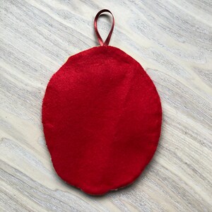 Puede incluir: Adorno ovalado de fieltro rojo con un lazo de cinta roja en la parte superior.