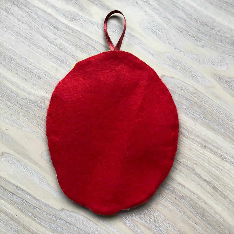 Puede incluir: Adorno ovalado de fieltro rojo con un lazo de cinta roja en la parte superior.