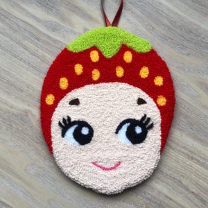 Puede incluir: Una fresa de fieltro rojo y verde con una cara sonriente, ojos negros y pesta&ntilde;as blancas. La fresa tiene semillas amarillas y est&aacute; colgada de una cinta roja.