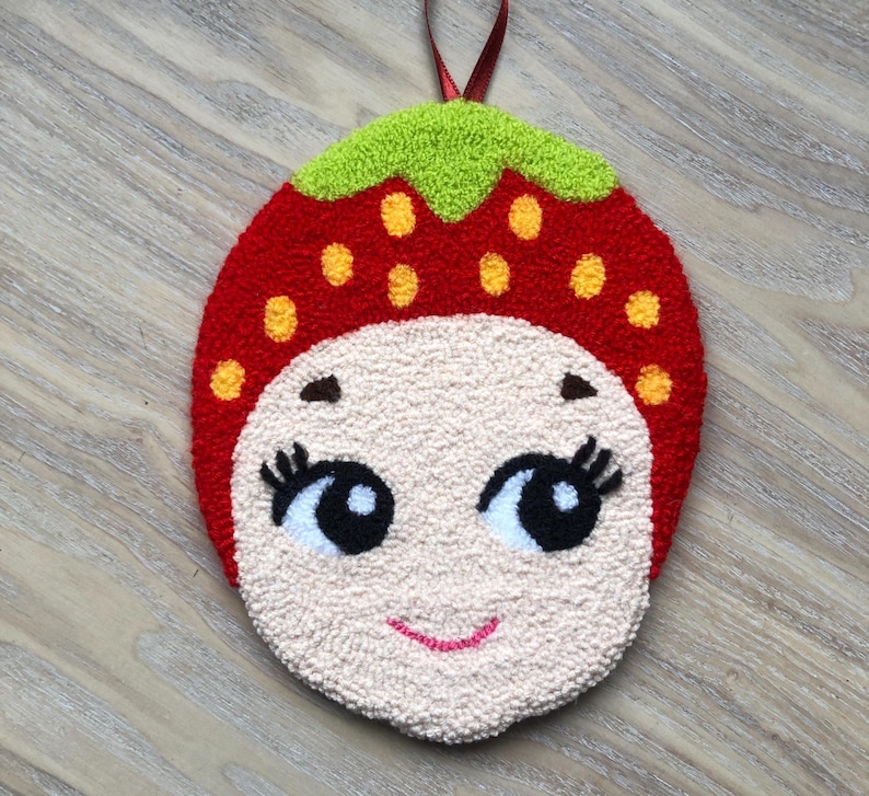 Puede incluir: Una fresa de fieltro rojo y verde con una cara sonriente, ojos negros y pesta&ntilde;as blancas. La fresa tiene semillas amarillas y est&aacute; colgada de una cinta roja.