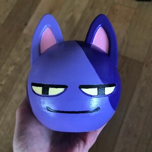 May include: A purple and blue cat figurine with a grumpy expression. The cat has yellow eyes and a black mouth.