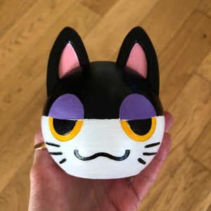 May include: A black and white cat figurine with purple eyelids and yellow eyes. The figurine has a white face with a black nose and whiskers.