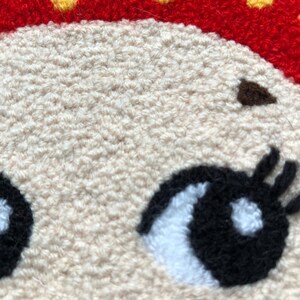 Puede incluir: Primer plano de una cara bordada blanca y esponjosa con ojos negros y pesta&ntilde;as. Los ojos tienen reflejos blancos. La cara es parte de un dise&ntilde;o bordado m&aacute;s grande.