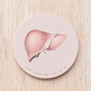 Puede incluir: Un posavasos redondo rosa con una ilustración dibujada a mano de un hígado y el texto "Life loves the liver of it".