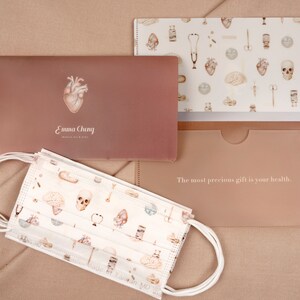 Pode incluir: Um conjunto de três pastas rosa e brancas com um tema de ilustração médica. As pastas têm um fundo branco com ilustrações de corações, cérebros, ossos e outros símbolos médicos. O texto "O presente mais precioso é a sua saúde." está impresso na parte inferior da pasta. As pastas são feitas por Emma Cheng, Medical Art & Gifts.