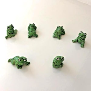 May include: Six small green ceramic frog figurines arranged in a group. Each frog features a distinct pose, including sitting, reclining, and resting positions, with painted black eyes and detailed textures on their bodies, showcasing varied expressions.