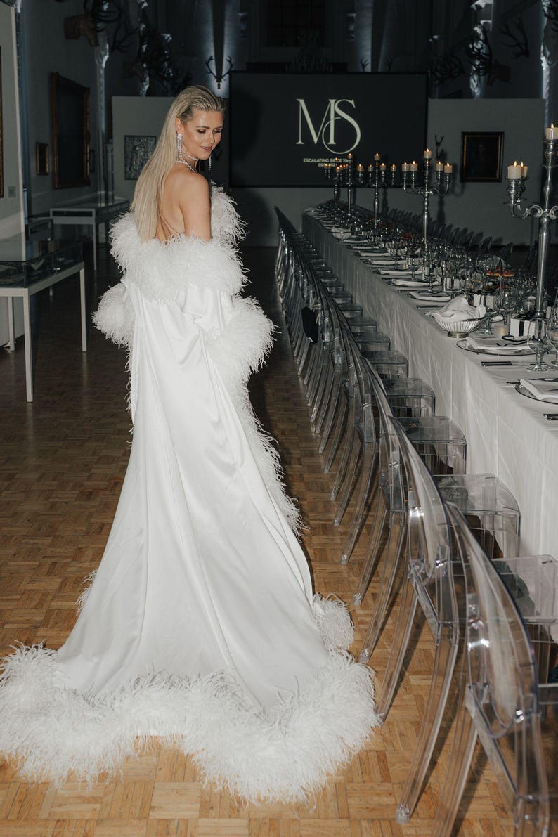 May include: A woman wearing a white satin gown with a feather trim. The gown is floor-length and has a plunging neckline. The woman is standing in a room with a long table set with white linens and clear chairs. A sign above the table reads 'MS Exclusive Suite Dinner'. 