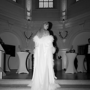 May include: A white feathered cloak with a satin lining. The cloak is worn by a person standing on a set of stairs in a room with large windows and white walls.