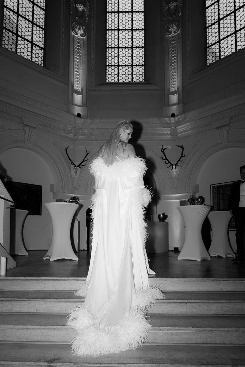 May include: A white feathered cloak with a satin lining. The cloak is worn by a person standing on a set of stairs in a room with large windows and white walls.