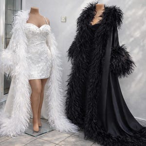 Puede incluir: Dos batas largas hasta el suelo con mangas largas y ribete de plumas en los bordes. Una bata es blanca y se combina con un minivestido blanco con escote coraz&oacute;n y detalles de encaje floral. La otra bata es negra y de aspecto satinado.