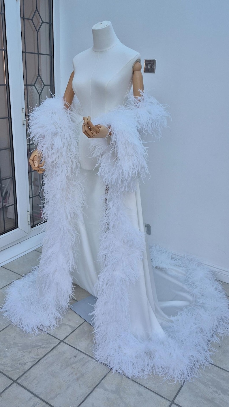 Puede incluir: Un vestido blanco hasta el suelo con un corpi&ntilde;o sin mangas y una falda larga y fluida. La prenda est&aacute; adornada con un extenso ribete de plumas blancas a lo largo de los bordes delanteros y el dobladillo, creando un aspecto voluminoso.