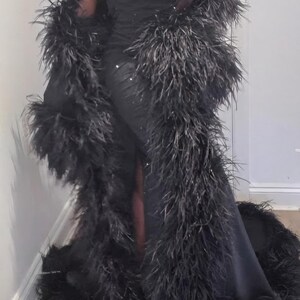 Puede incluir: Un vestido negro largo hasta el suelo con tirantes finos y una bata larga negra a juego con un borde de plumas densas y voluminosas a lo largo de los bordes delanteros y el dobladillo. La tela del vestido tiene un brillo sutil.