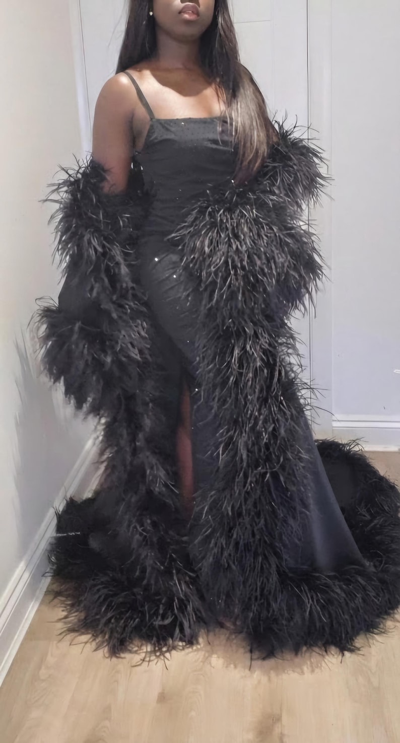 Puede incluir: Un vestido negro largo hasta el suelo con tirantes finos y una bata larga negra a juego con un borde de plumas densas y voluminosas a lo largo de los bordes delanteros y el dobladillo. La tela del vestido tiene un brillo sutil.