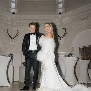 May include: A couple in a formal setting. The woman is wearing a white gown with a feather trim and the man is wearing a black tuxedo with a black bow tie.