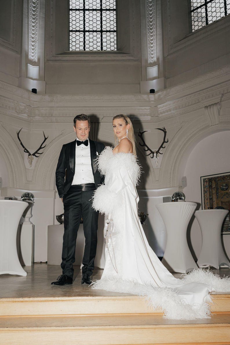 May include: A couple in a formal setting. The woman is wearing a white gown with a feather trim and the man is wearing a black tuxedo with a black bow tie.