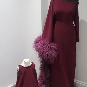 Könnte beinhalten: Zwei burgunderfarbene Kleider mit Federnbesatz. Das größere Kleid hat einen langen Ärmel und das kleinere Kleid hat keine Ärmel. Beide Kleider haben einen burgunderfarbenen Federnbesatz am Saum und einen Ärmel.